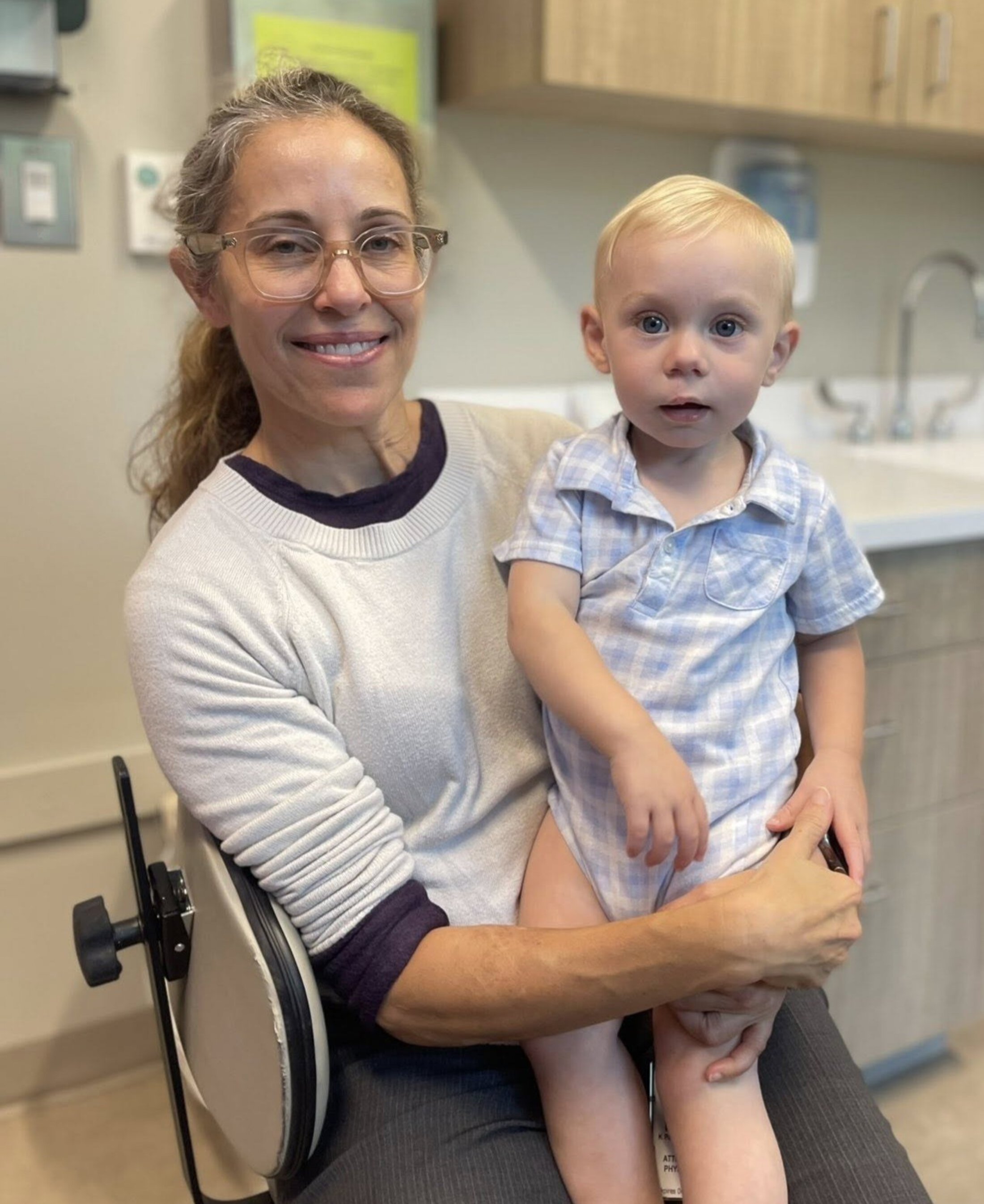 Photograph showing an adult seated on a chair holding a young child in a medical examination room. Background includes wooden cabinets, a sink, and medical supplies, indicating a healthcare setting.