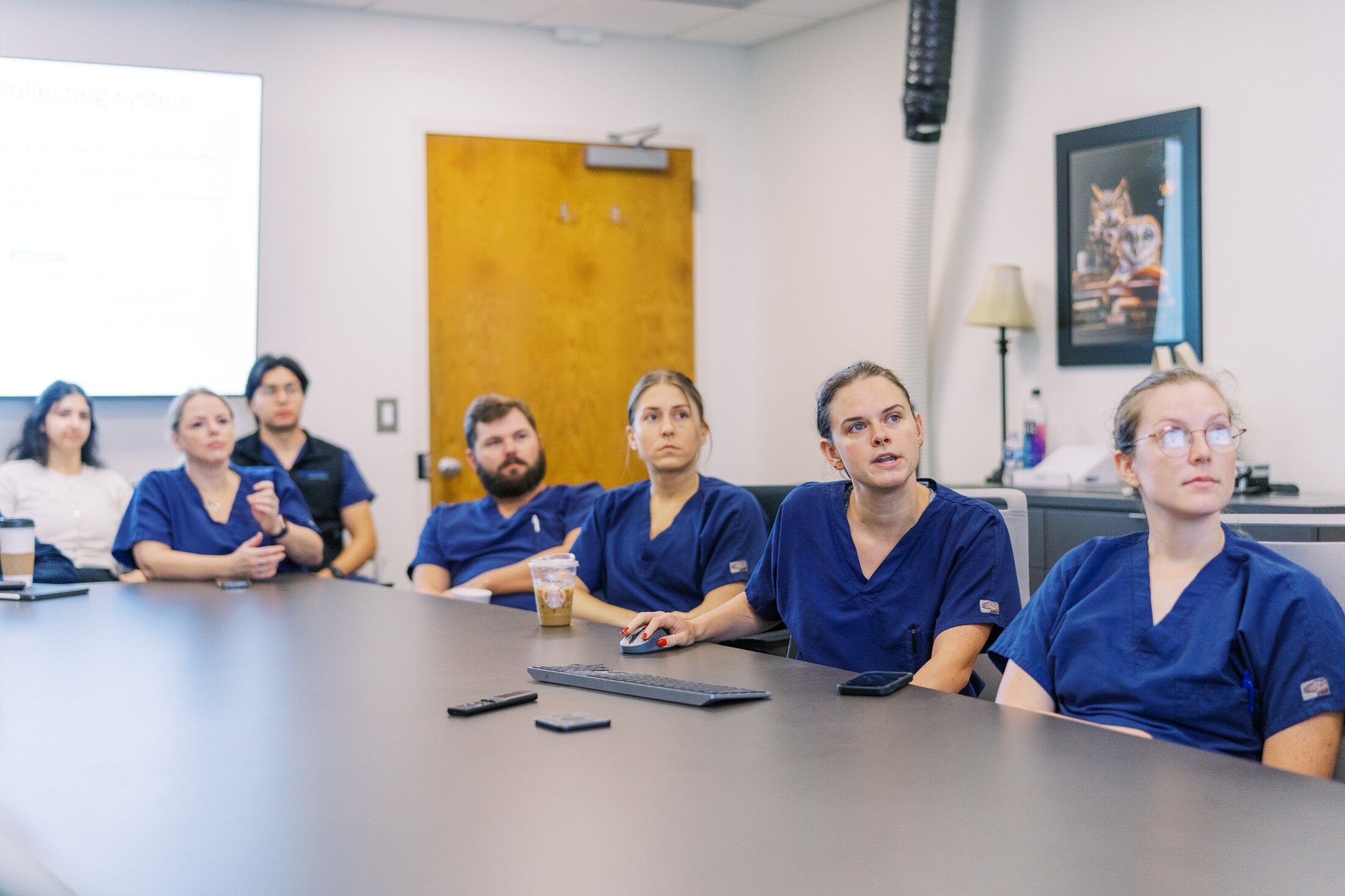 A resident clicks through a presentation she is discussing with the peers in scrubs seated around the conference table with her.