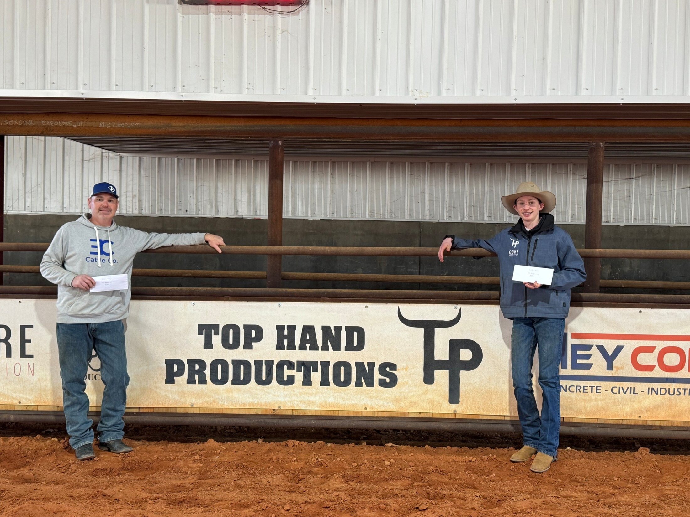 A man and teen boy pose casually against the railing of a rodeo arena, holding envelopes with their winnings.