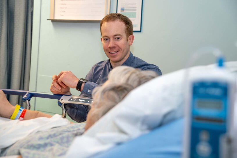 A man sits bedside in a hospital room with an elderly female patient.