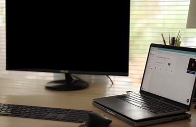 Laptop on a desk next to a blank external monitor, with a keyboard and mouse in front and window light coming through blinds.