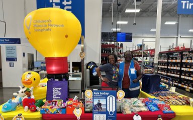 Two Costco employees stand arm-in-arm behind a CMNH fundraising booth for MUSC Children's Health. The booth contains bright colored toys, snacks and a table-sized inflatable red and yellow hot air balloon with the Children's Miracle Network Hospitals logo.