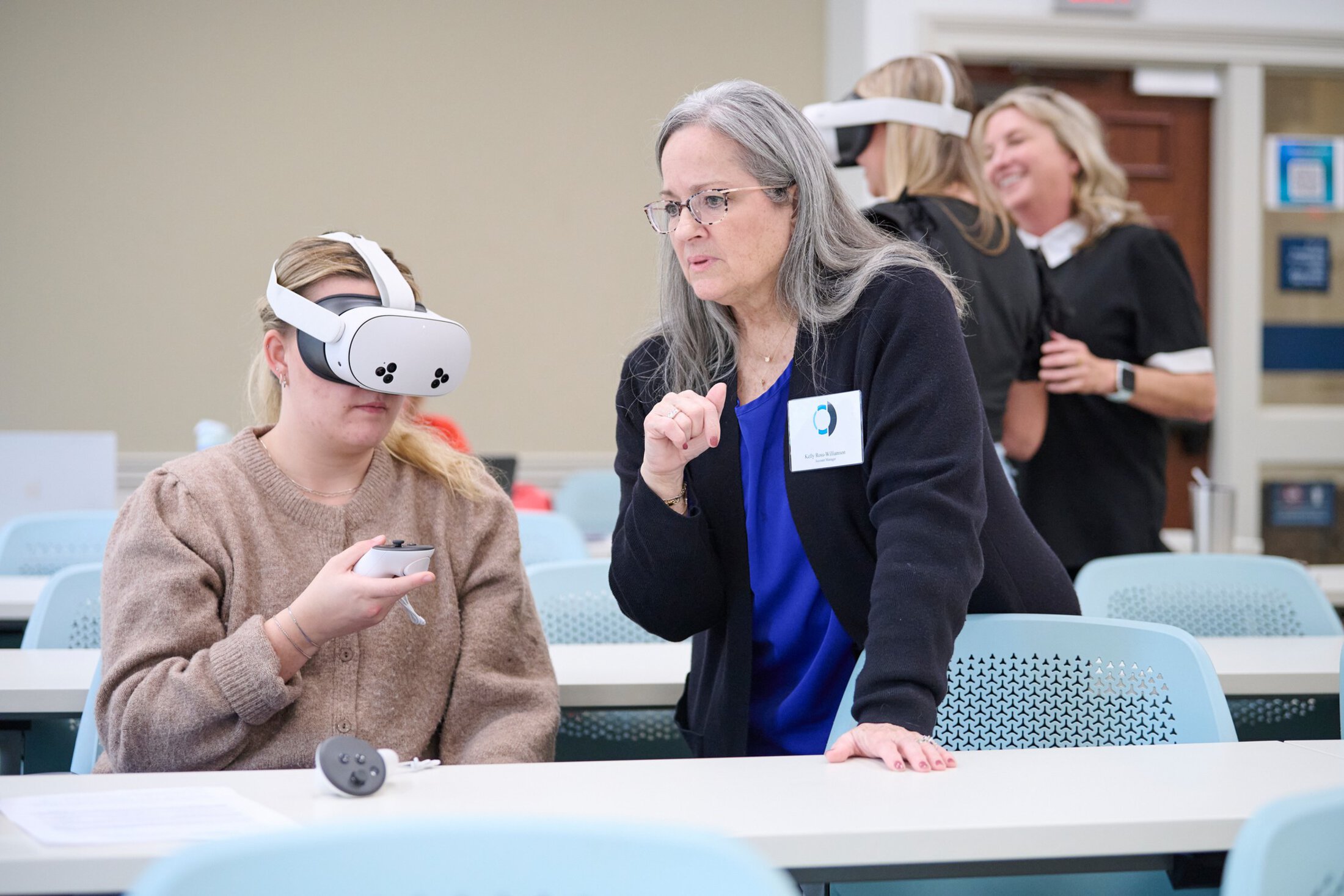A nursing student wearing a virtual reality headset holds a controller while seated in a classroom as an instructor stands beside her offering guidance during a cognitive impairment care simulation. Other participants using VR headsets are visible in the background.
