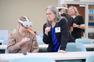 A nursing student wearing a virtual reality headset holds a controller while seated in a classroom as an instructor stands beside her offering guidance during a cognitive impairment care simulation. Other participants using VR headsets are visible in the background.