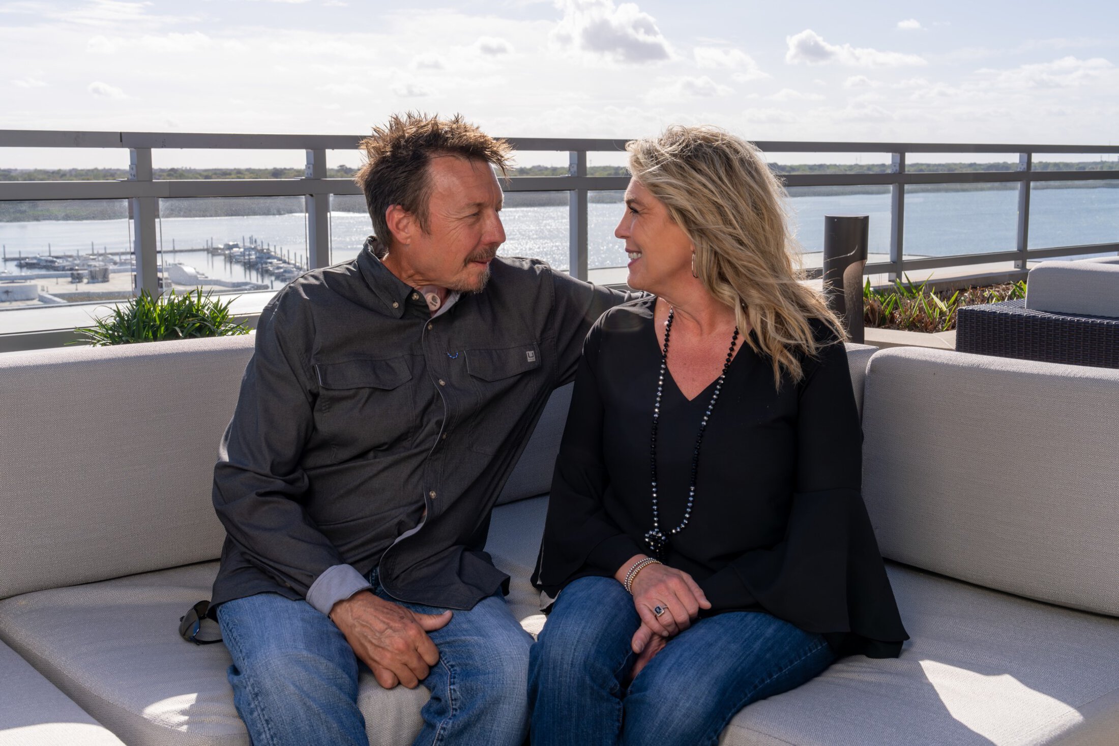 A couple sit on a patio sofa on a rooftop deck overlooking a river and marina and grin at each other.