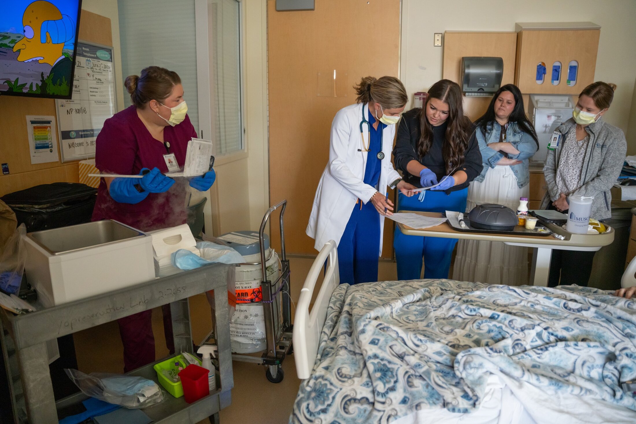 Four women, a doctor and nurses, cluster around a bedside table in a hospital room and look over a printout while off to the side a woman wearing cryogenic gloves holds a plastic container recently pulled from a liquid nitrogen container as ice fog wafts beneath her hands. A scene from The Simpsons plays on the TV mounted on the wall.
