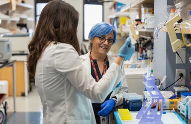 Two women in a lab. One is facing the camera. She has blue and white hair and is wearing round glasses. The other woman's face is covered by long brown hair. They are both wearing white lab coats.