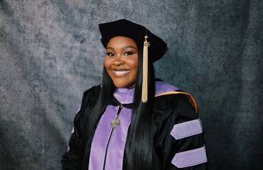 Smiling woman with long dark hair wearing a black cap with a gold tassel and the number 26 hanging from it. She's wearing a black graduation robe with purple accents.