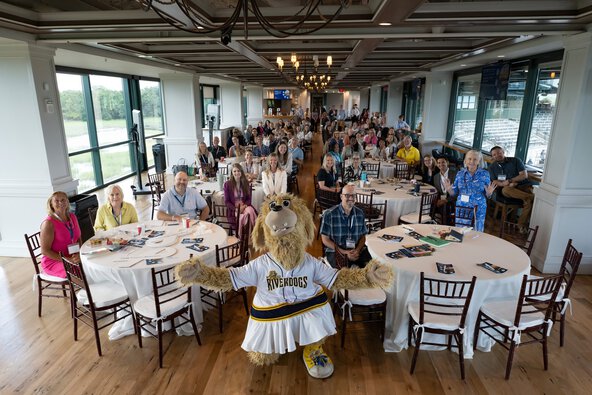 A large event space full of people seated at tables with the mascot for the Charleston Riverdogs baseball team kneeling in front.