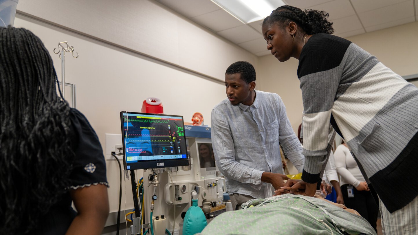 Two students stare intently at a monitor as one student performs compressions on a medical manikin while another uses a bag valve mask to deliver oxygen.