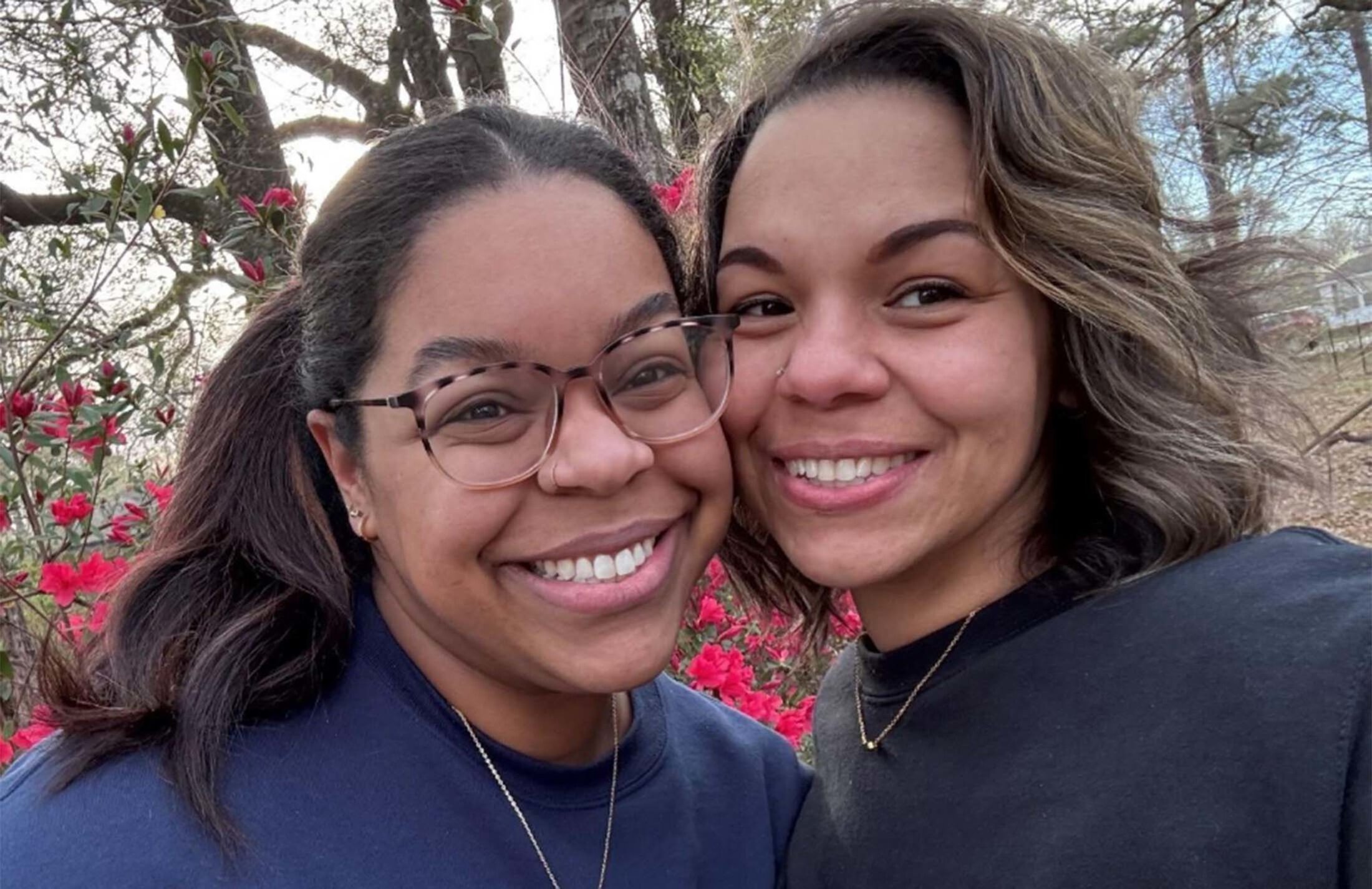 Two women smile as they lean into each other. The woman on the left has her hair in a ponytail and is wearing glasses. The woman on the right has shoulder length hair.