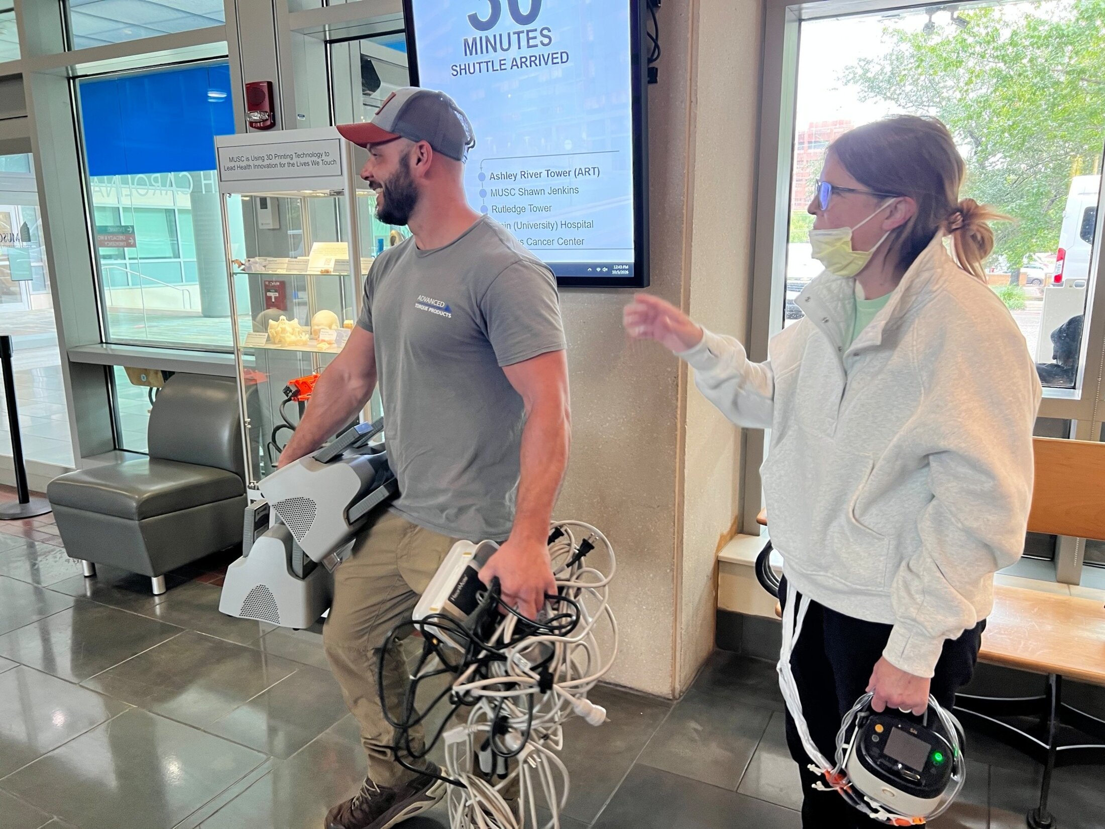 A man wearing a ballcap, t-shirt and khakis and a woman wearing a surgical mask, sweatshirt and dark pants, carry wired equipement through a lobby.