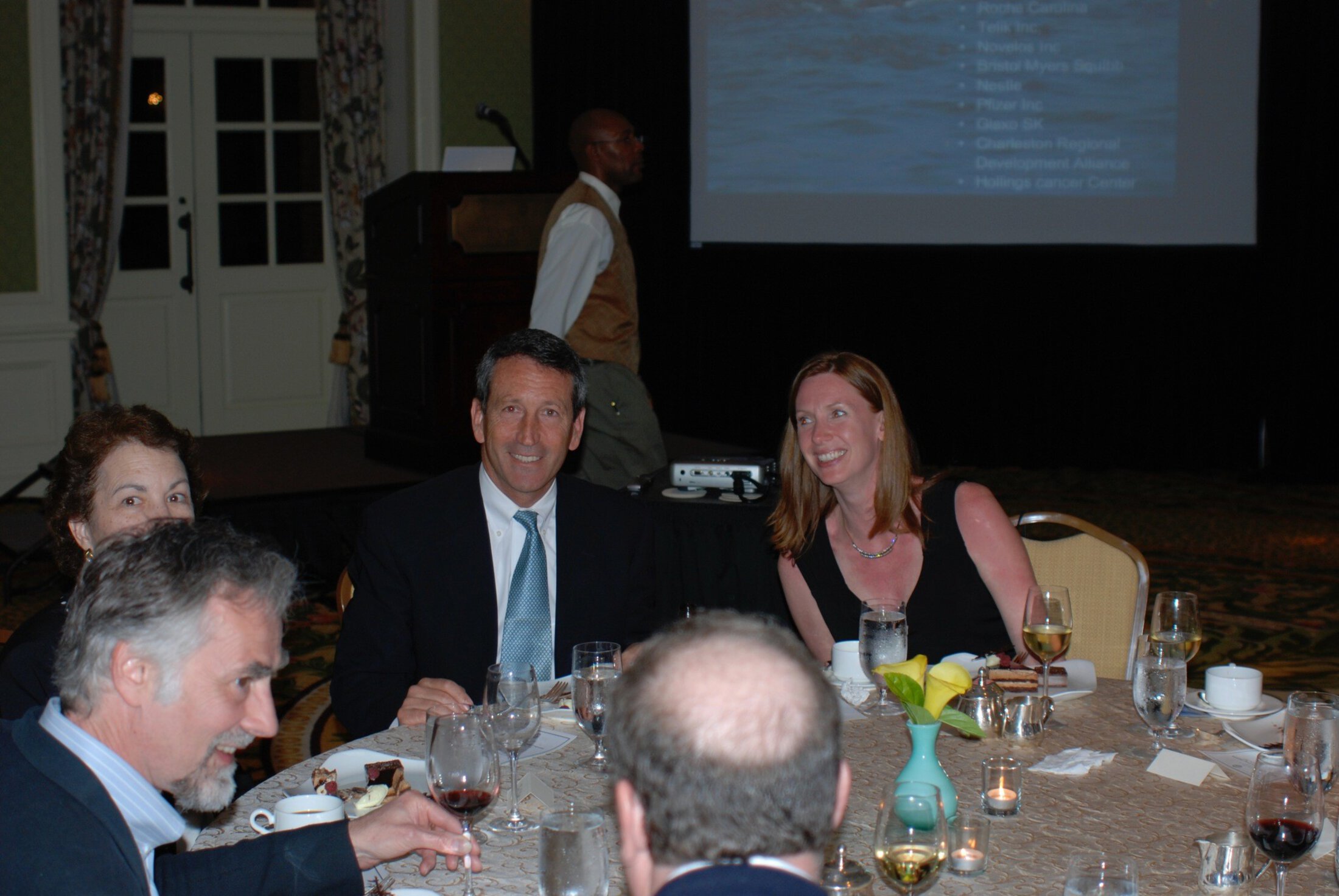 Former South Carolina Governor Mark Sanford, wearing a dark jacket, white shirt and necktie, smiles while seated at a table. A woman to the right smiles as she leans toward him. There are three other people at the table.