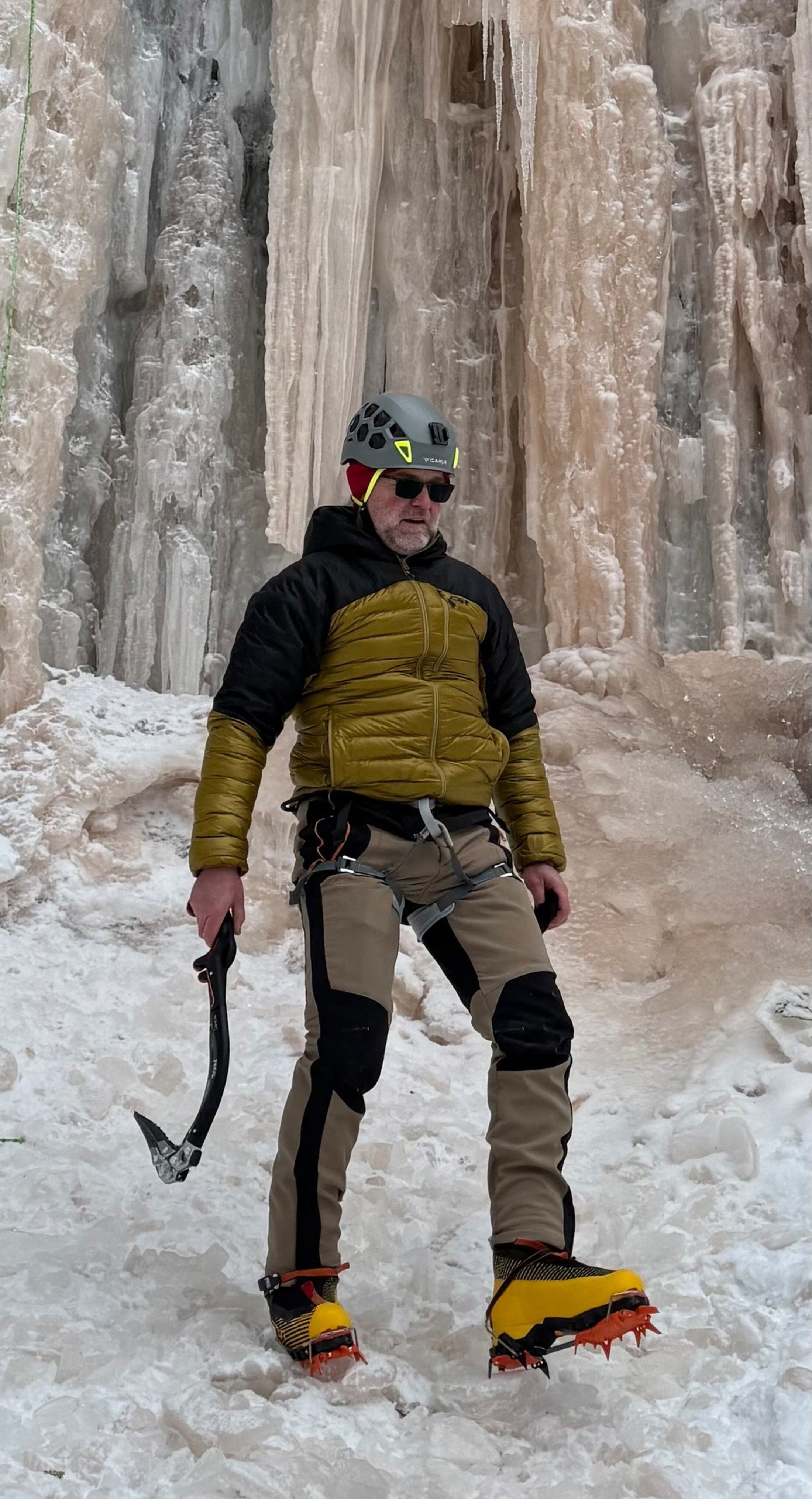 A man wearing spiky ice boots walks in front of an ice wall.