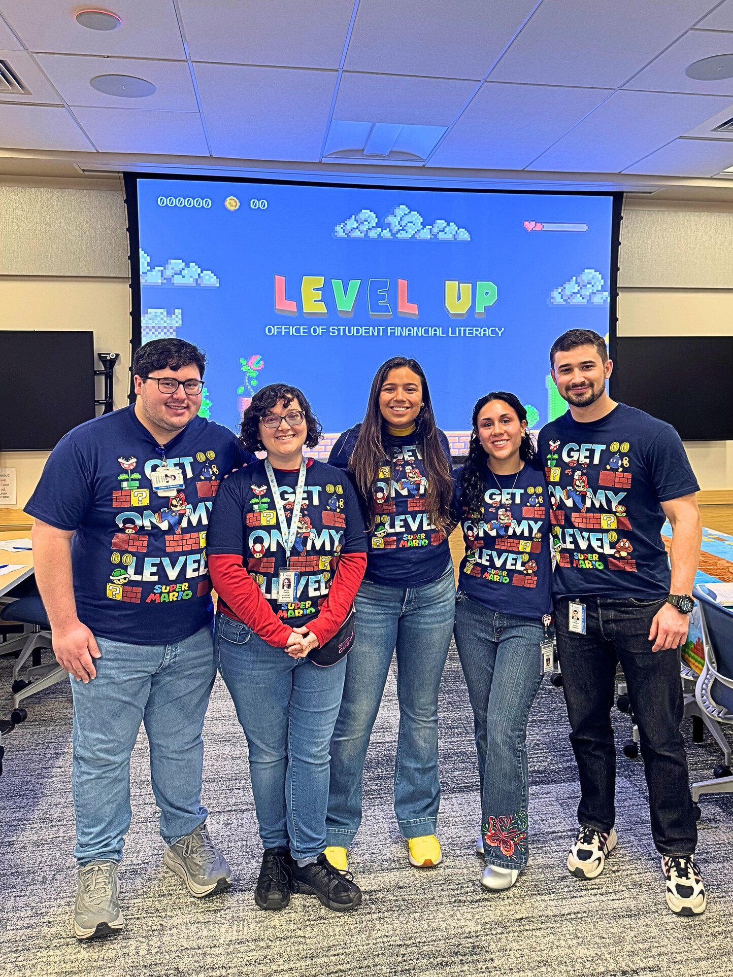 Five student mentors wearing “Get on My Level” shirts stand together in front of a Level Up financial literacy presentation screen.