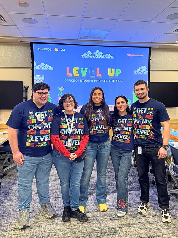 Five student mentors wearing “Get on My Level” shirts stand together in front of a Level Up financial literacy presentation screen.