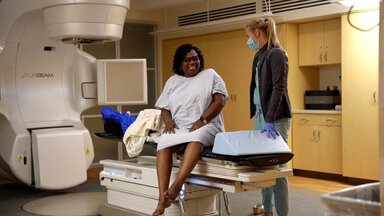 A woman in a patient gown sits on the radiation therapy table.