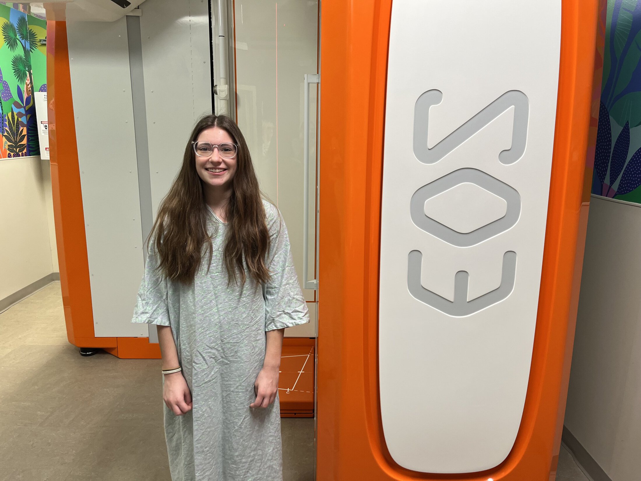 A young woman with long brown hair and glasses, wearing a hospital gown, smiles while standing next to a large orange and white EOS imaging machine. A tropical mural is partially visible in the background.