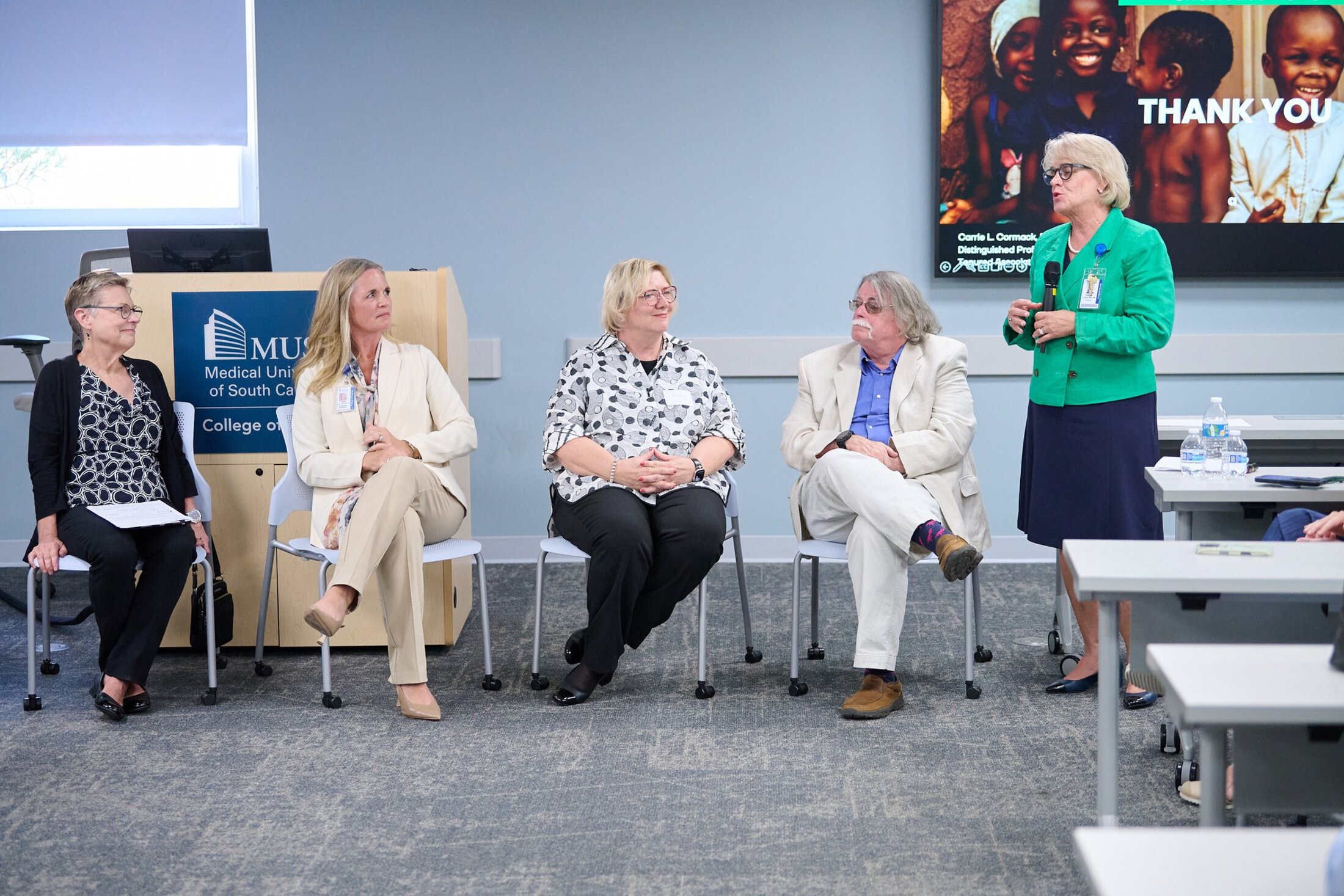 Four in chairs, a woman is standing on the right and holding a microphone while she speaks. The seated people are three women and one man. They are all wearing business casual clothing.