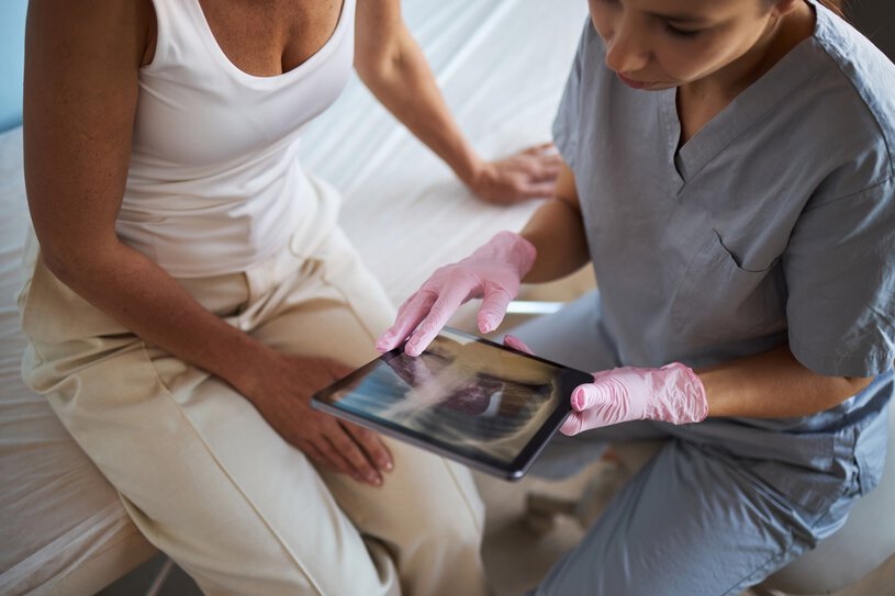 Middle aged Caucasian woman sitting beside young adult Caucasian female doctor wearing medical gloves, showing chest X-ray image on digital tablet, during cancer consultation
