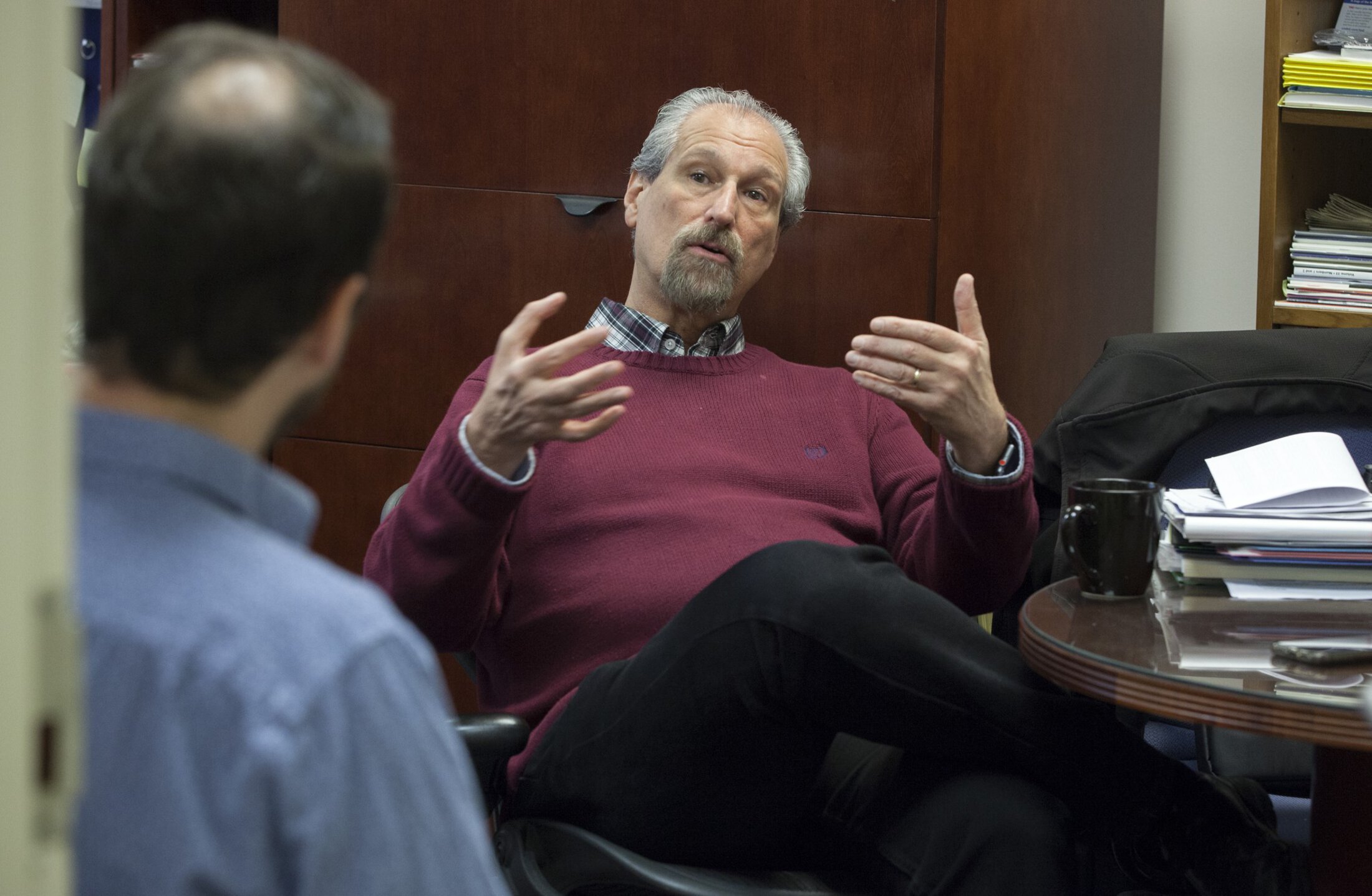 A man is seated wearing a reddish sweater and plaid button down shirt. He is gesturing with his hands. He has gray hair and a goatee. There is a cup of coffee on a table beside him.