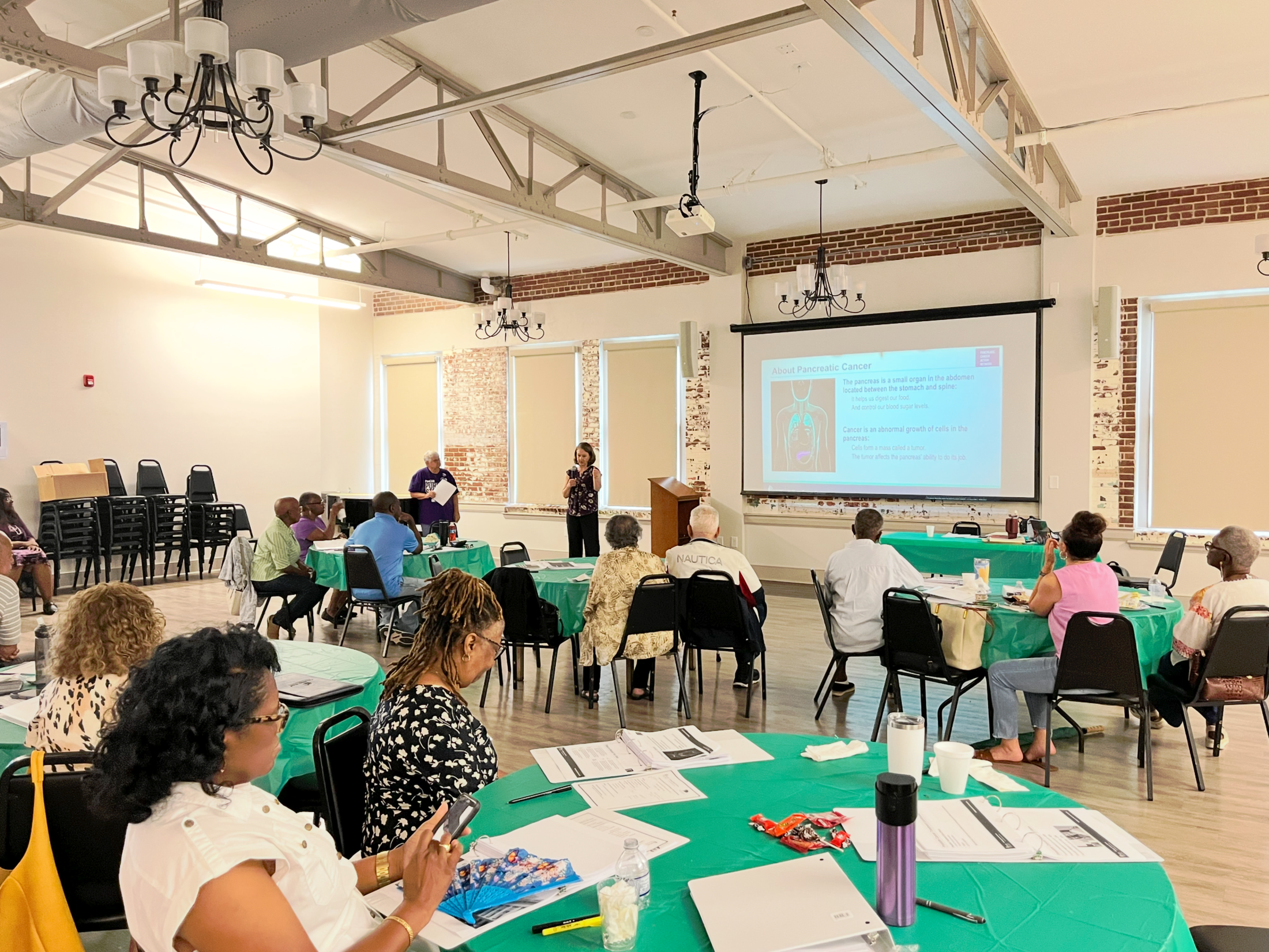 People sit around banquet tables scattered with workbooks, pens and cups and listen as a woman speaks with a pancreatic cancer information slide on the screen behind her.