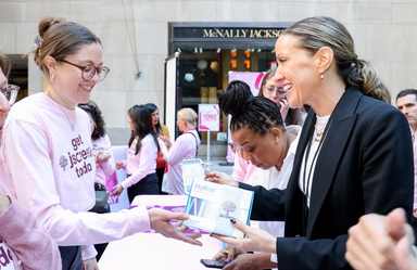 A smiling woman hands a cancer screening packet to another woman at an event. Others gather in the background.