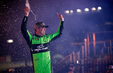 A man wearing a green andn black jumpsuit and a ball cap raises his arms. He's holding what looks like a bottle of water and smiling.