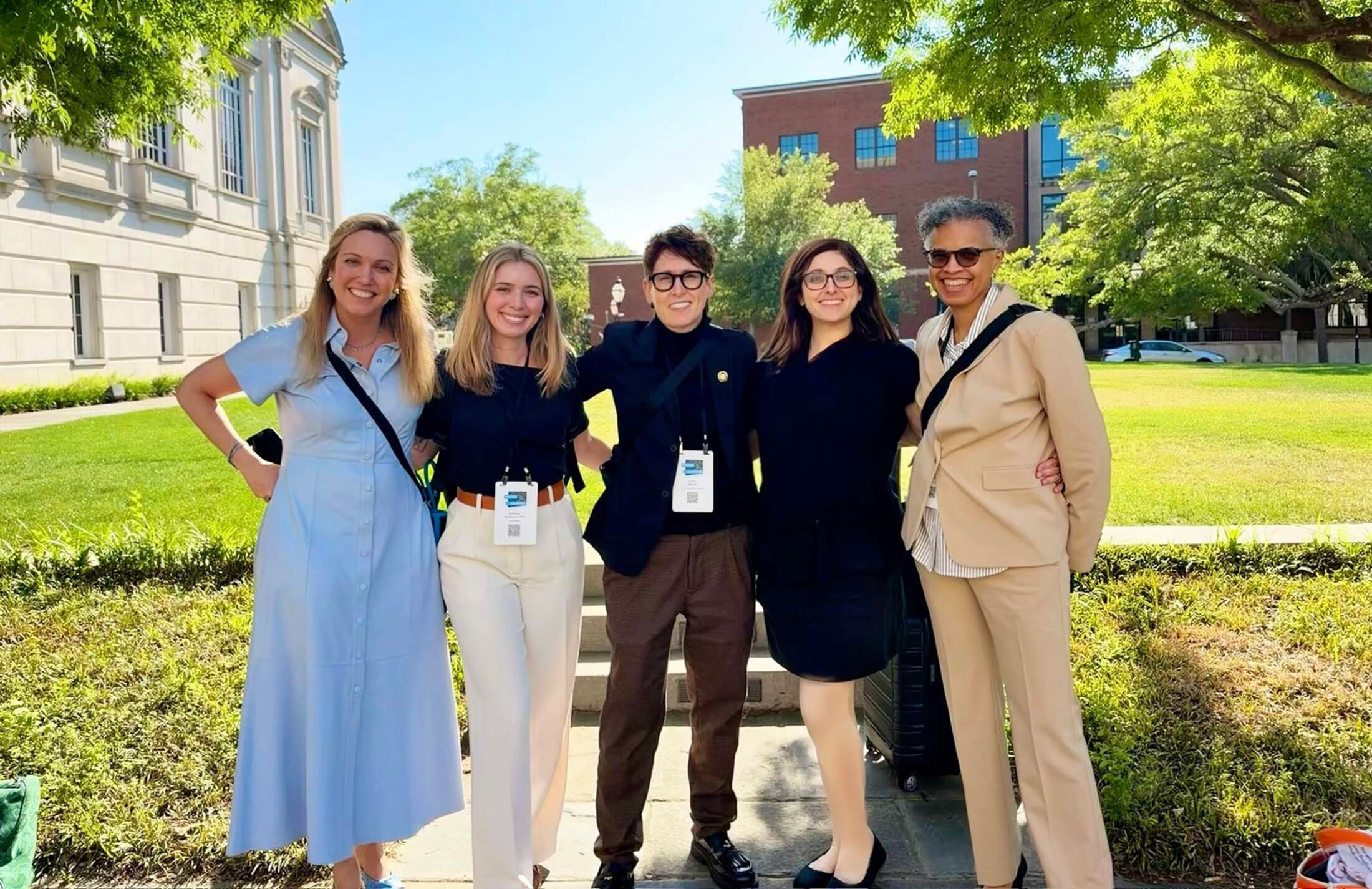 Five women stand together outside with two large buildings behind them. The two women on the far left are blonde with long hair. The woman in the middle has short dark hair and is wearing glasses. The two women on the right are also wearing glasses. All are wearing business casual type clothing.