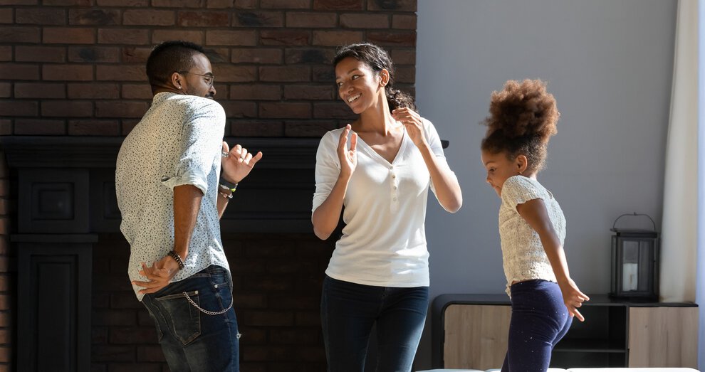 Happy young african american family with little daughter dancing together in living room, overjoyed biracial parents and small girl child tenants have fun celebrate moving to new home or apartment