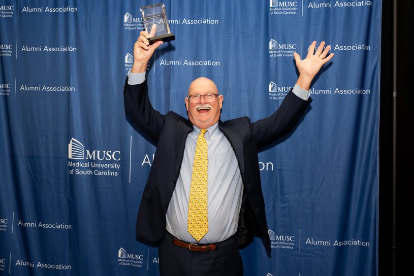 A smiling man in a suit raises an award trophy with both hands in celebration while posing in front of a Medical University of South Carolina Alumni Association backdrop.