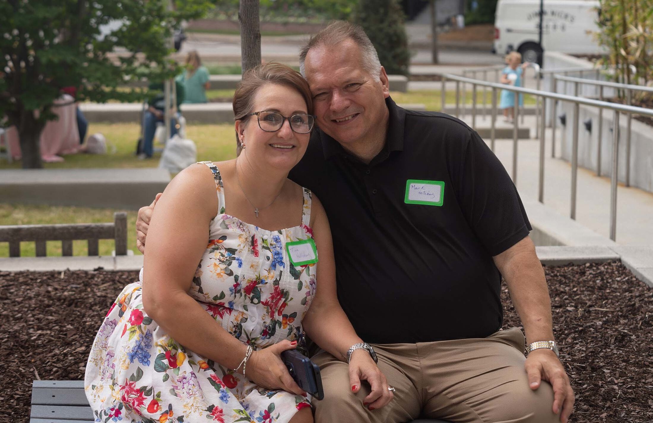A woman wearing a floral dress and eyeglasses leans into a man wearing a black polo shirt and khakis. They are outside next to a rail and grass is behind them.