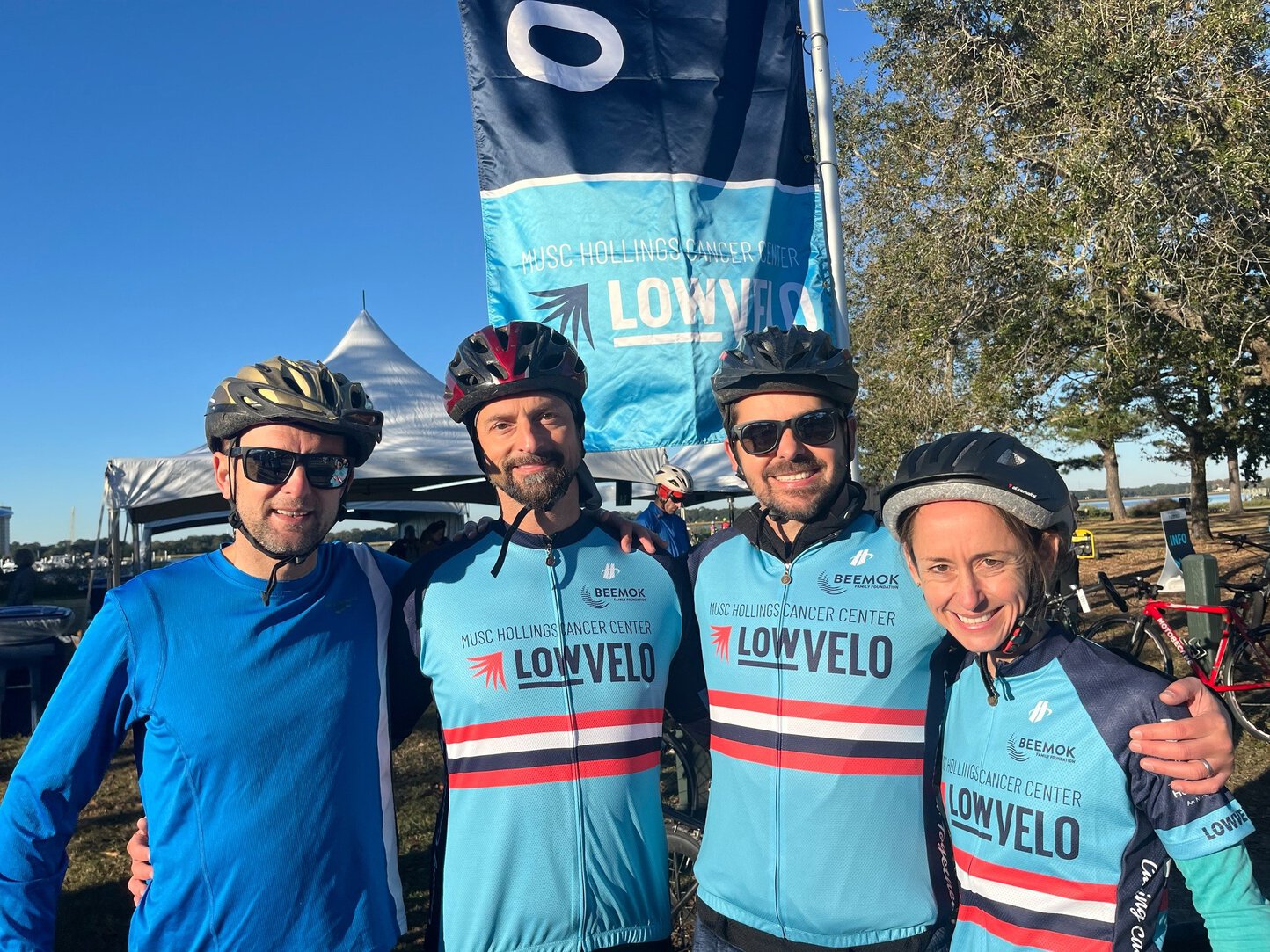 Four adult siblings, all wearing bike helmets, three wearing LOWVELO jerseys, gather under a LOWVELO banner flapping in the breeze.