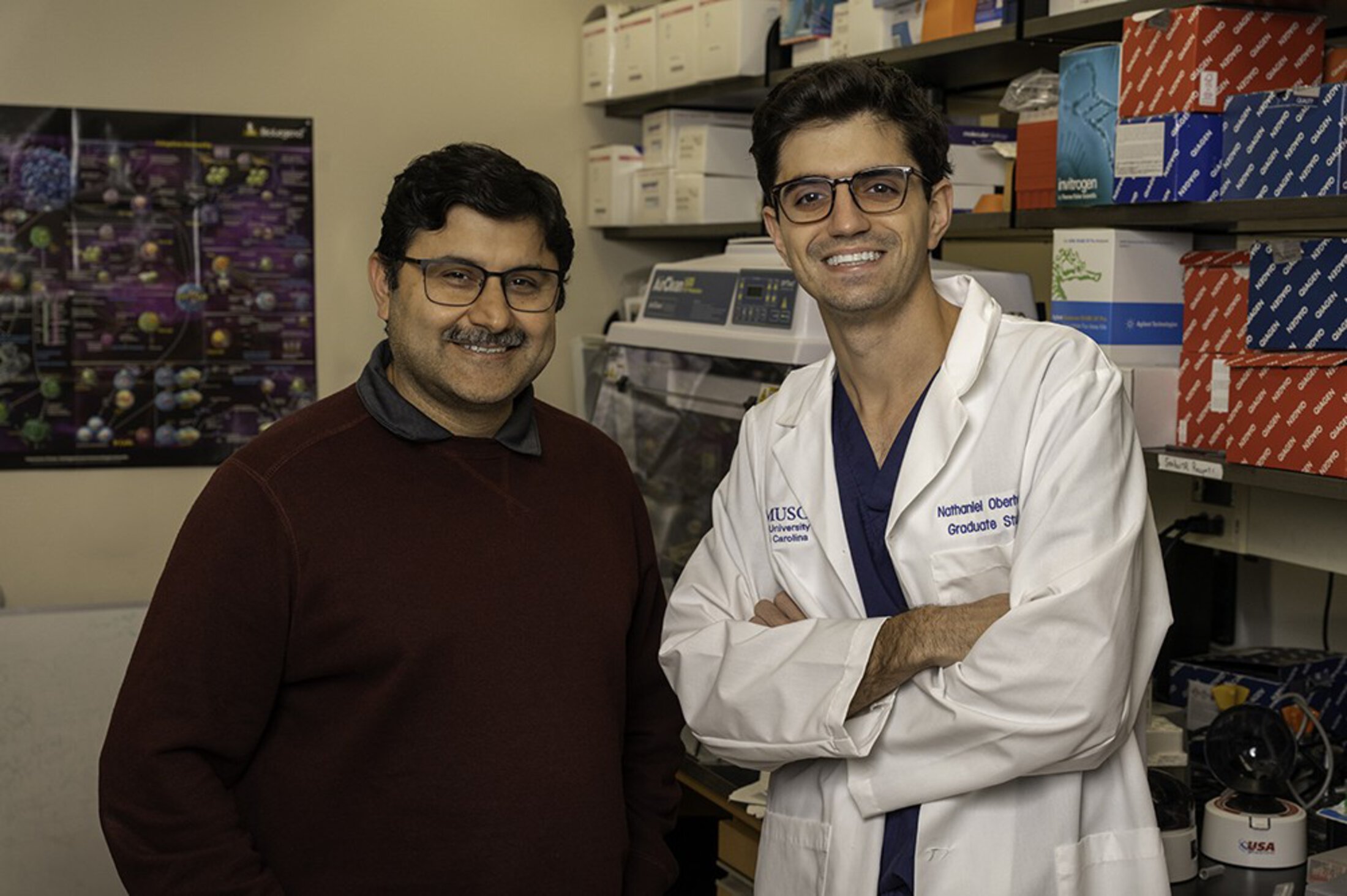 A man in casual business attire and a man in scrubs and a white lab coat that says 'graduate student' in a lab.