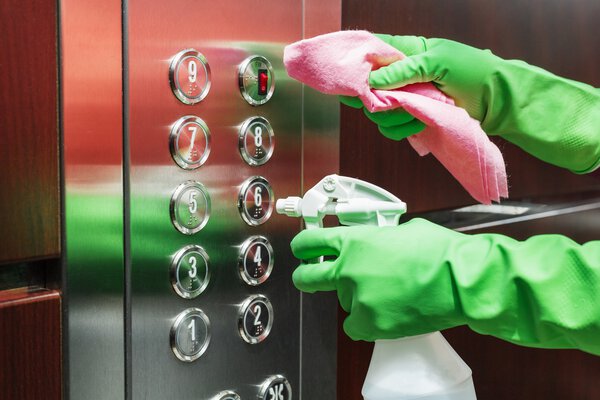 A person uses a disinfectant spray and a wet wipe to clean the elevator button control panel