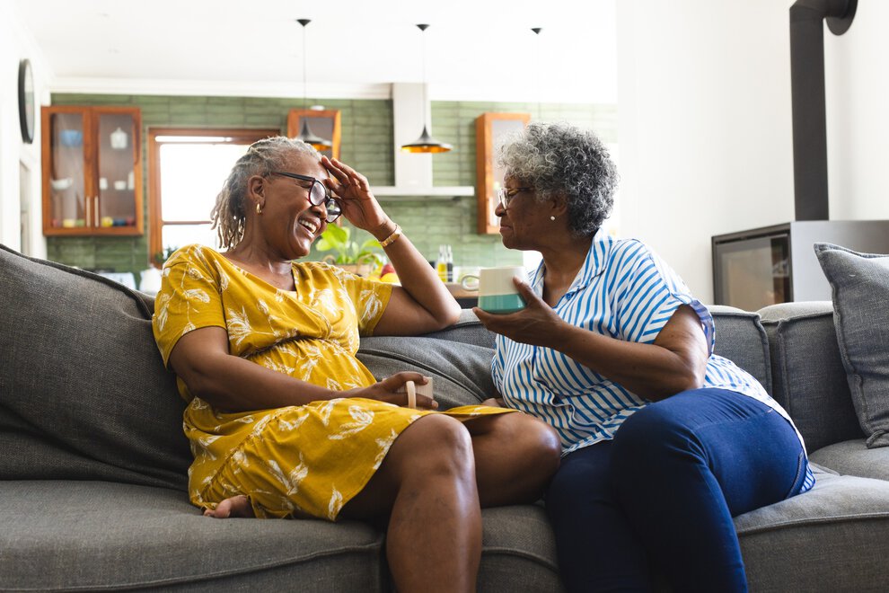 Two older women laugh over coffee shared on a comfy sofa.