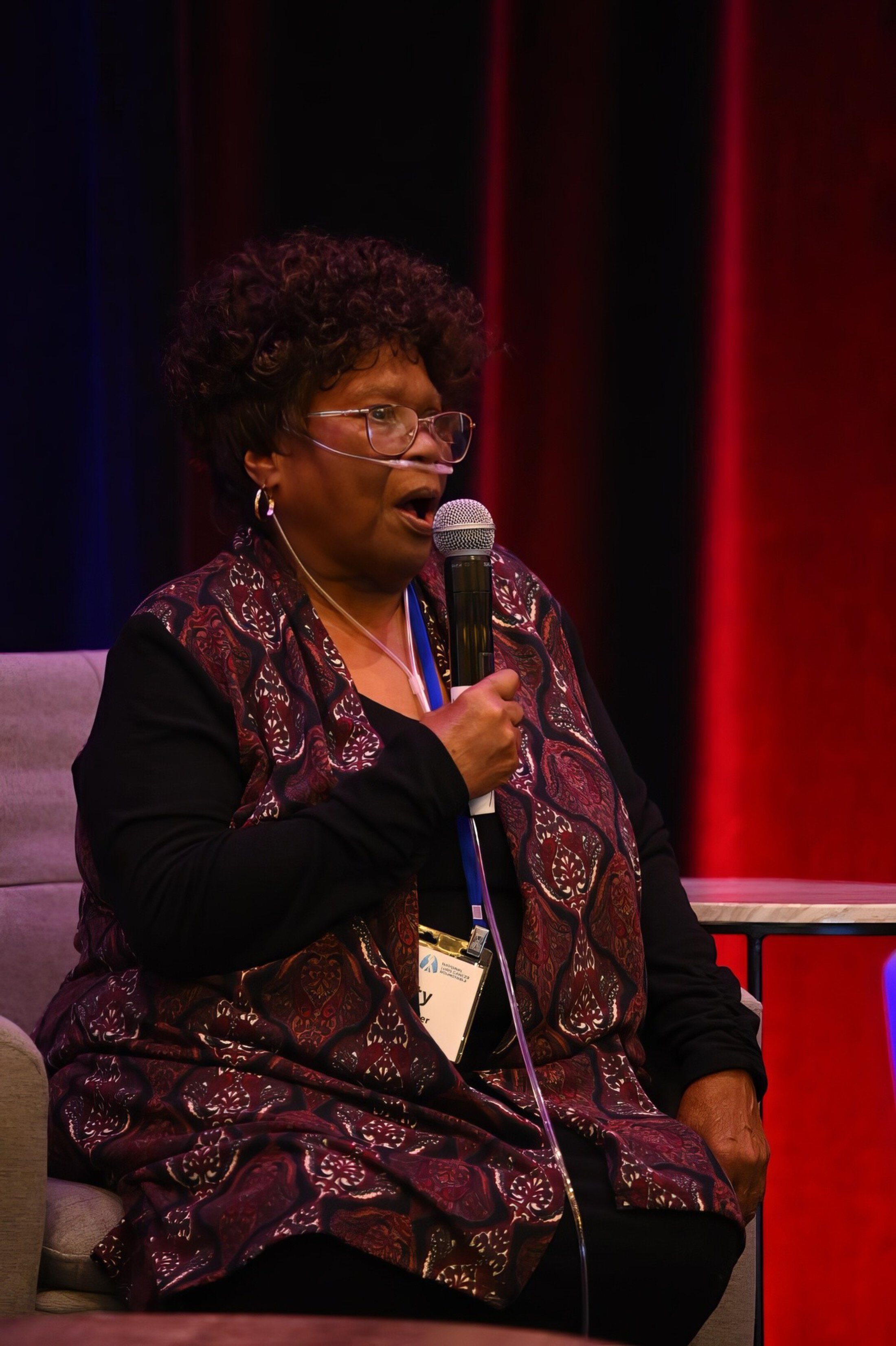 A woman wearing an oxygen tube speaks into a microphone while seated on stage.
