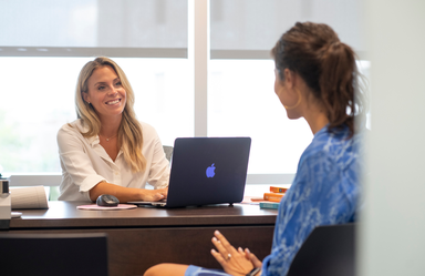 two women sit across from each other at a desk. one woman smiles at the other in front of an open laptop.