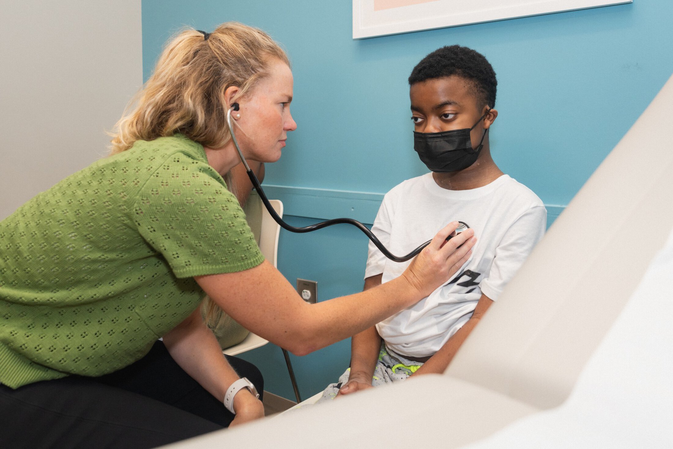 A woman with blonde hair wearing a green top has a stethoscope. She's checking the heart of a boy who is wearing a black surgical mask and a white t-shirt.