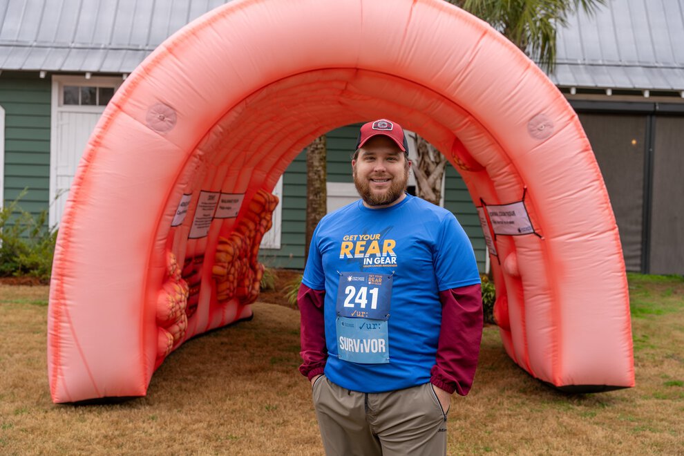 A man wearing a 'Get your rear in gear' shirt and a race bib stands in front of a giant inflatable colon.