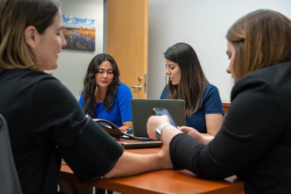 Four women sit at a table in an office talking in pairs.