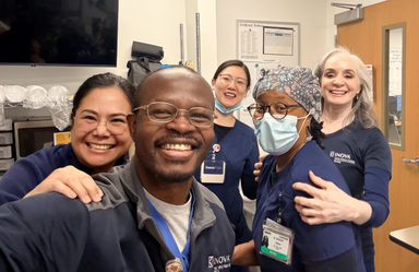 Five ECMO/perfusion professionals smiling together in a clinical workspace, smiling, wearing scrubs and ID badges.