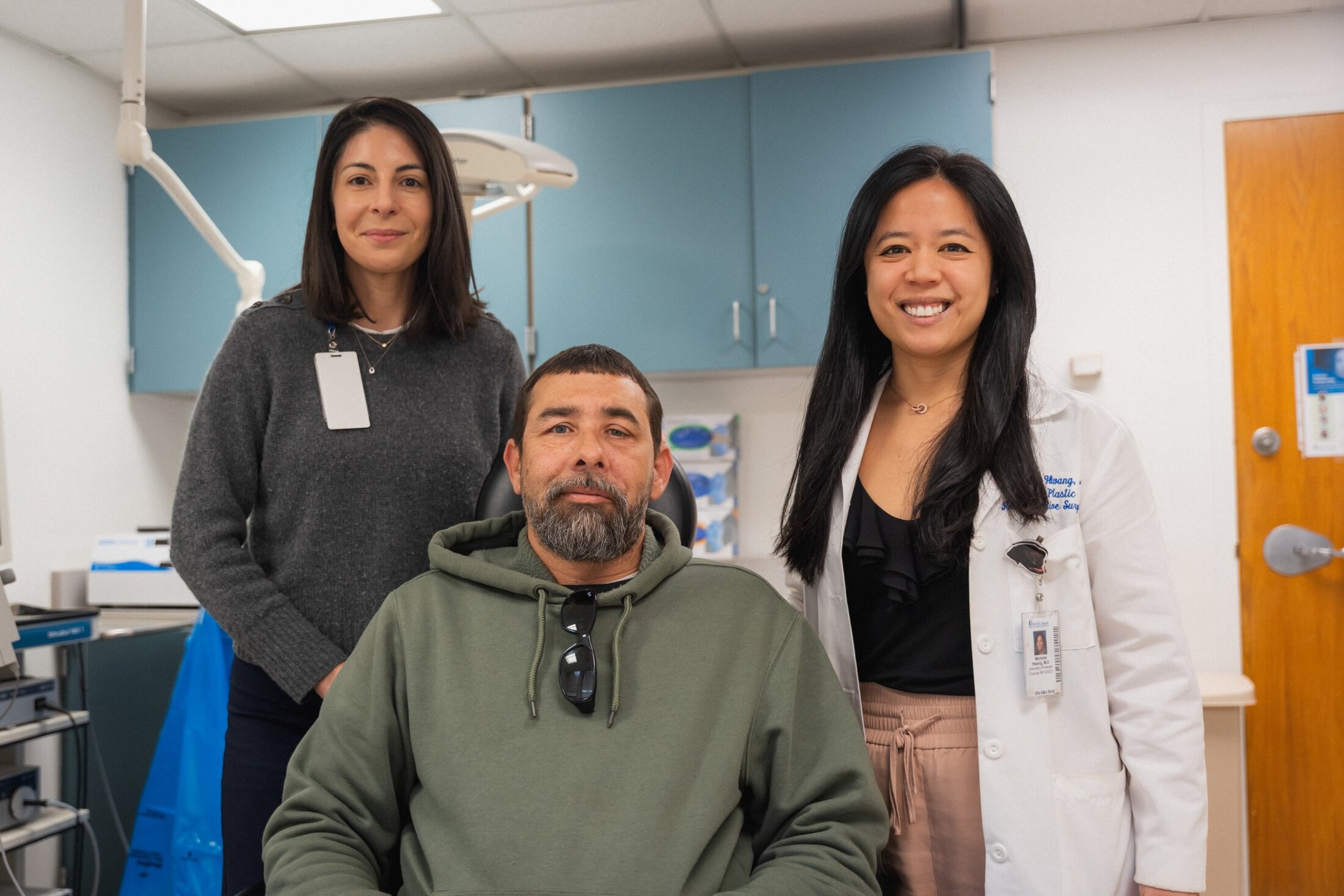Two women stand behind a seated man. They are in a medical clinic. The woman on the left is wearing a grey sweater and has a tag hanging from her colllar. The woman on the right is wearing a white doctors' coat. You can see the words 