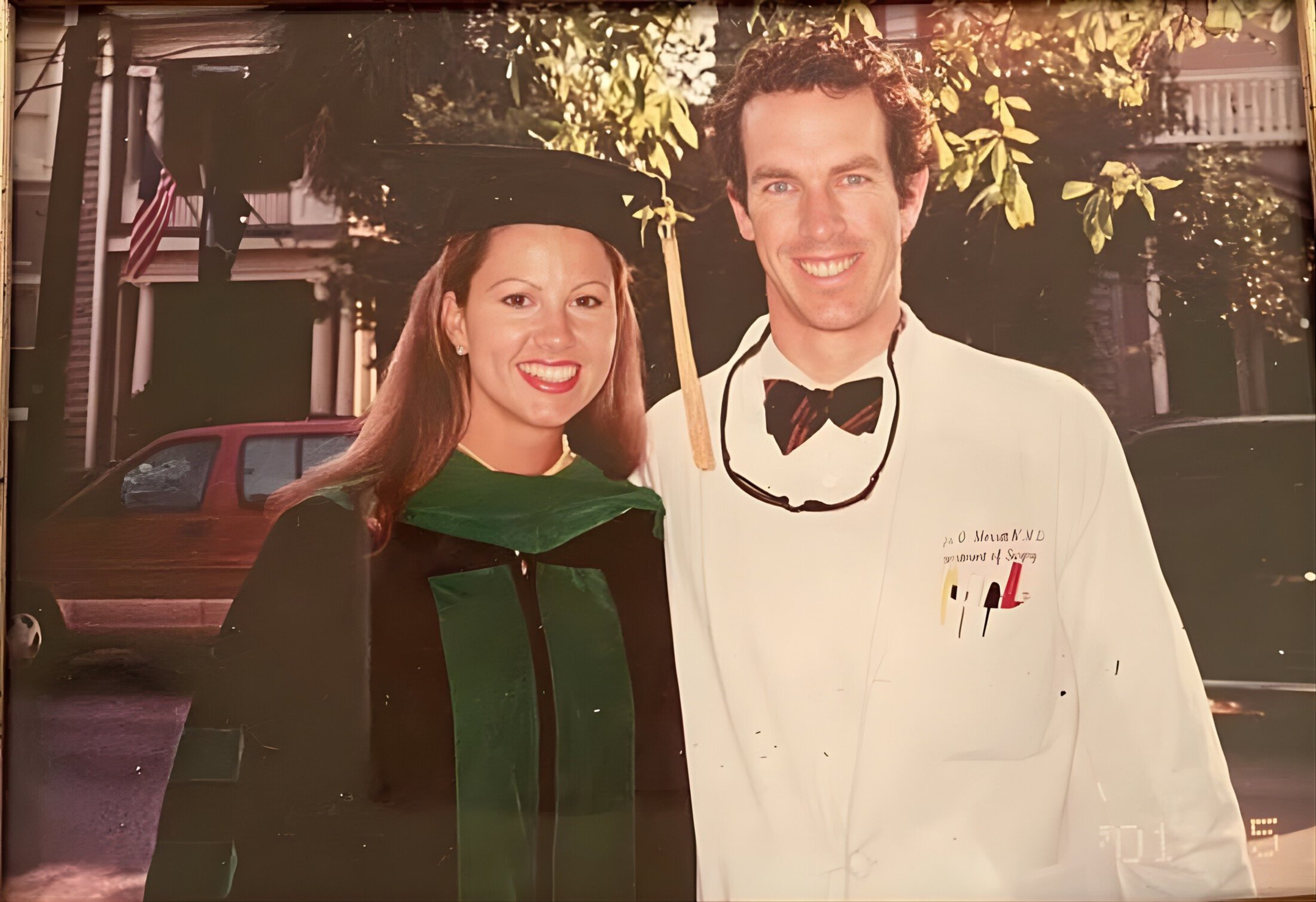 Graduation photo featuring a smiling woman in a green and black academic gown and cap, and a man in a white coat with a bow tie. Background shows a tree-lined street with a house and American flags, conveying a joyful and celebratory atmosphere.