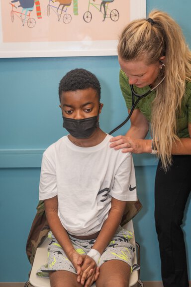 A child wearing a black surgical mask, a white t-shirt and patterned shorts is seated as a woman with blonde hair and a stethoscope checks his heart.