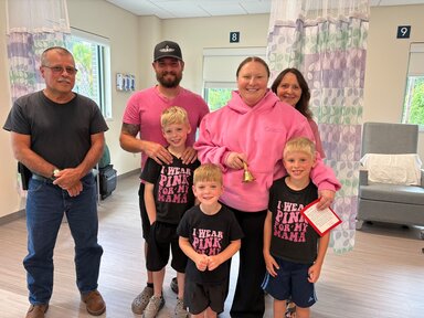 A woman in a pink sweatshirt holds a handbell as she is surrounded by family, including her husband in a pink shirt, three little boys in shirts that say 'I wear pink for my mama' and her parents in a cancer infusion center.