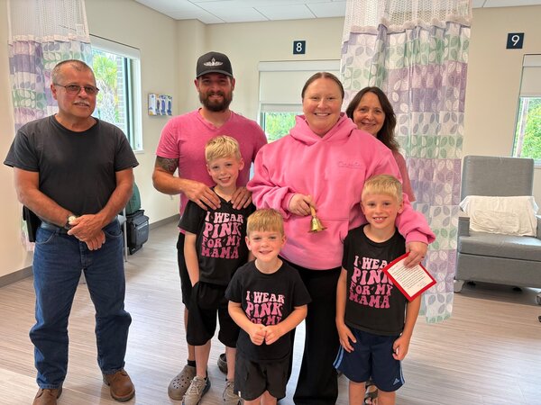 A woman in a pink sweatshirt holds a handbell as she is surrounded by family, including her husband in a pink shirt, three little boys in shirts that say 'I wear pink for my mama' and her parents in a cancer infusion center.