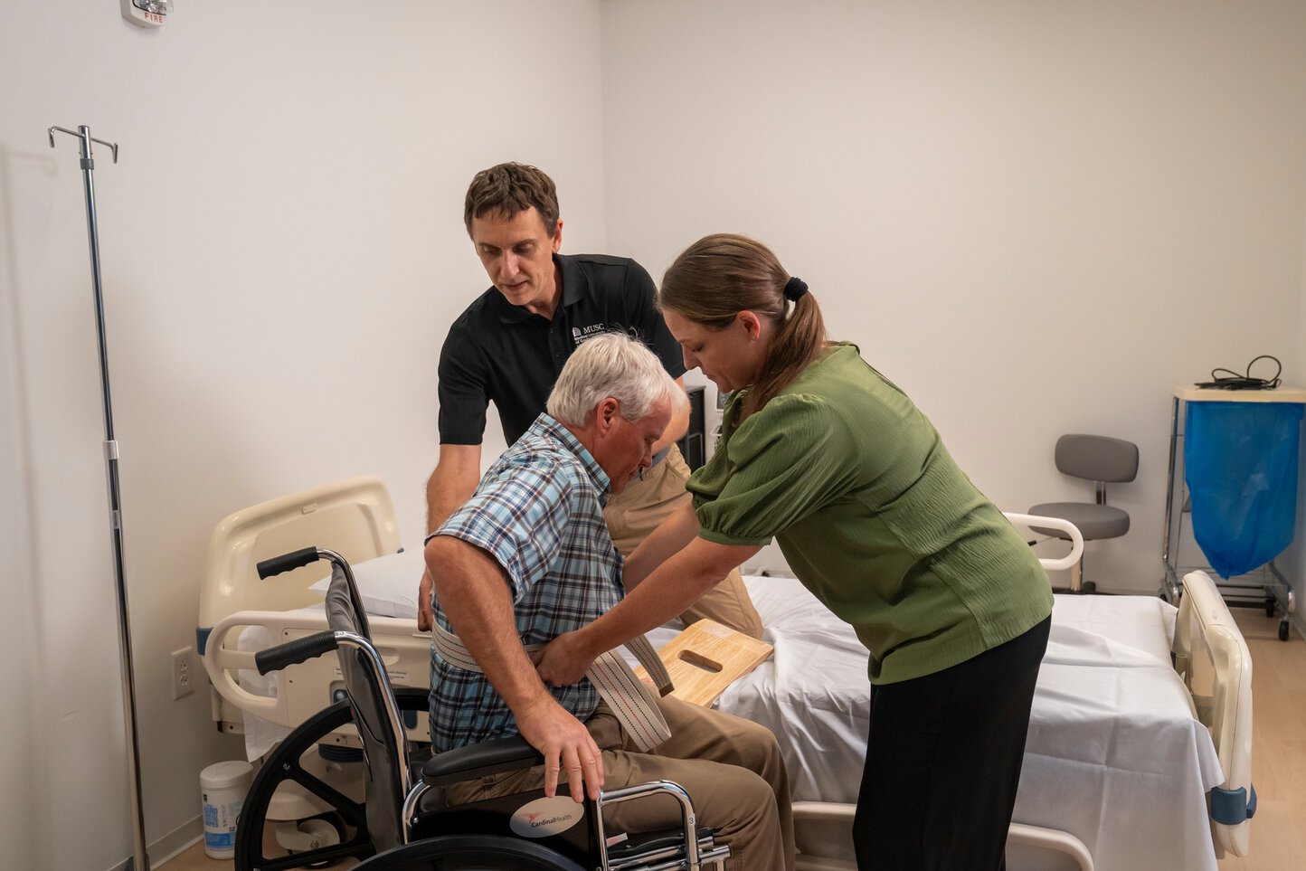 A man guides a woman in the safe transfer of a man from a wheelchair to a hospital bed.