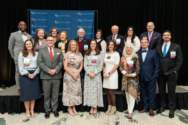 A group of about 18 professionally dressed adults stand and sit together on a stage in front of a blue MUSC Alumni Association backdrop, posing for a formal event photo. Several people wear name badges and award ribbons, and one woman in the front row holds a glass award.