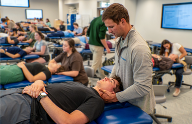 A man performs a neck adjustment on a prone man in a PT lab setting.